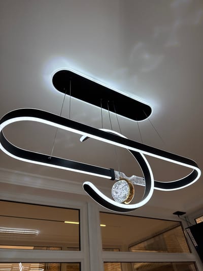 This is a close-up, low-angle indoor photograph of a modern, multi-tiered black chandelier with integrated LED lighting. The chandelier features an elongated, flattened oval base attached to the ceiling, from which two large, curved black arms extend downwards, each outlined with a continuous strip of white LED light. A smaller, ornate circular pendant hangs from the lower arm. The pendant consists of a gold-toned ring surrounding a clear, faceted orb filled with what appears to be small, shimmering particles. The background is a plain, light-colored ceiling, with some subtle reflections visible. Below the chandelier, the top of a white-trimmed window frame with tinted glass is visible, reflecting ambient light and the chandelier itself. The photograph is taken from a perspective that emphasizes the dramatic curves and illumination of the fixture. There are no people or signs of activity in the image. The location is identified as Baş-Bulak, Kyrgyzstan, however, no visual elements in the image confirm this specific geographical location.
