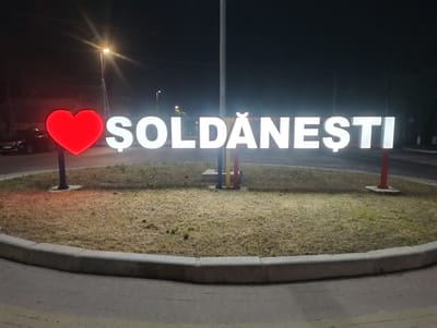 The image shows a large, illuminated sign spelling "ȘOLDĂNEȘTI" in white, block letters with a red heart to the left of the text. The sign is positioned on a circular patch of dry grass, bordered by a concrete curb and a paved area. The scene is at night, with streetlights visible in the background, suggesting an outdoor urban or semi-urban environment. The lighting from the sign is bright, illuminating the immediate surroundings. The location appears to be an entrance to the city of Șoldănești, Moldova, given the text on the sign. No people are present in the frame.