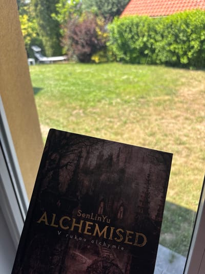 The image shows a book titled "ALCHEMISED" by SenLinYu, with the subtitle "V rukou alchymie" in Czech, placed on a windowsill. The book cover features a dark, gothic-style illustration of a forest and architecture. Outside the window, a sunlit green lawn is visible, with bushes and a red-tiled roof of a house in the background. The overall scene suggests an indoor view looking out onto a residential garden on a sunny day. The location is specified as Červená Píska, Czechia.