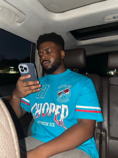 A medium shot shows a man with dark skin, short dark hair, and a beard, sleeping while holding a mobile phone in a car. He is wearing a light blue short-sleeved shirt with white and red stripes on the sleeves, and grey shorts. The text "CUSTOM WOL..." and "CHOPPER CUSTOM BIK" are visible on the shirt. The car's interior has grey roof upholstery, and the man is leaning against the interior. The car has dark brown leather seats. Outside the window, city lights are seen, suggesting a nighttime environment. The specific location of Jiru, Nigeria, cannot be determined from the image alone.