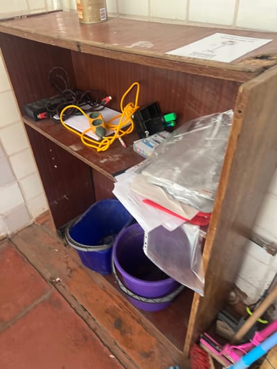 The image shows a cluttered wooden shelf unit in what appears to be a storage area or utility room. On the top shelf, a metal can is visible. The second shelf from the top contains a black power strip with a coiled yellow cord, a black electronic device, and a white piece of paper with a diagram. To the right, there is a black object with green markers and a clear plastic bag filled with papers. The lower shelf holds two buckets, one blue and one purple. The area below the shelves is visible, showing a tiled floor and a wooden base. To the right of the shelf unit, cleaning tools including brooms and mops are leaning against the wall. The overall environment appears somewhat disorganized and worn. The location is identified as Mlyniv, Ukraine, though no specific landmarks are visible in the image to confirm this. There are no people present in the image.