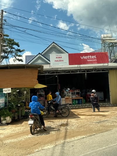 The image depicts a street scene in Tu Póh, Vietnam. In the foreground, a person wearing a blue hooded jacket is riding a motorcycle with license plate 49-P1 04378. To the right, another person wearing a dark striped robe and a white helmet is walking. In the background, there are buildings with businesses. A red banner with "Viettel telecom" is visible on one of the buildings. A sign on another building reads "TẬP HÓA KIM NGÂN". The environment appears to be a sunny day with a blue sky and scattered clouds. The road is unpaved and appears to be made of dirt and gravel. There are power lines overhead.