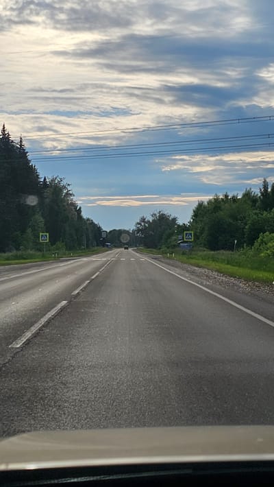 The image depicts a paved road, likely a highway or a main road, in Solnechnyy, Russia. The road has multiple lanes and is bordered by dense trees on both sides. The sky is partly cloudy with a mix of white and blue hues. There are power lines running horizontally across the upper portion of the image. A few cars are visible on the road, including one in the distance. There are also road signs present, including a yellow pedestrian crossing sign on the left side of the road. The overall scene suggests a rural or suburban environment outside of a major city.