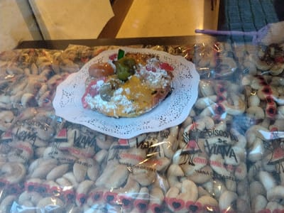 A bakery display case features a ring-shaped cake dusted with powdered sugar and decorated with candied fruits, resting on a white lace doily. Surrounding it are numerous packages of biscuits. The packages are transparent, revealing crescent-shaped cookies, some with heart-shaped decorations. The text on the cookie packages reads "Biscoitos de Viana" and "Confeitaria Viana do Castelo." The image suggests a selection of traditional Portuguese pastries available for purchase.