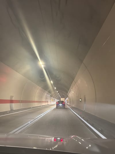 The image shows the interior of a tunnel, likely a road tunnel. The perspective is from the driver's seat of a vehicle, looking forward. The tunnel is illuminated by overhead lights, which create streaks of light in the image due to motion blur. The walls of the tunnel are curved and appear to be made of concrete, with a light-colored finish. There is a distinctive horizontal stripe, red on the left side of the tunnel and extending into the distance.
Two cars are visible in front of the viewpoint vehicle. The closest car is dark-colored and seen from behind, its taillights illuminated. Further ahead, another dark-colored SUV is visible, also from the rear. The road surface appears to be asphalt with white lane markings and small red markers along the side. The overall impression is of a modern, well-maintained tunnel.
The location is specified as Đurmani, Montenegro. Given that Đurmani is a coastal town, it's plausible this is a road tunnel designed to facilitate travel through mountainous terrain or beneath a body of water, common in such regions. The tunnel is designed for vehicular traffic, indicated by the presence of multiple cars and road markings. The lighting and structure suggest it is a functional piece of infrastructure for transportation.