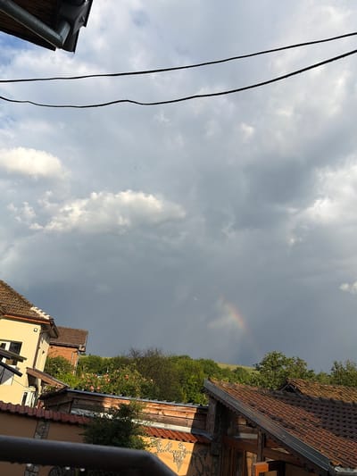 The image depicts a cloudy sky in Kozlovets, Bulgaria, with a faint rainbow visible. Overhead power lines are visible in the left foreground. The roofs of buildings, including a tile roof and a yellow-painted building, line the bottom of the frame. Green foliage, including trees and bushes, fills the mid-ground between the buildings and the sky.