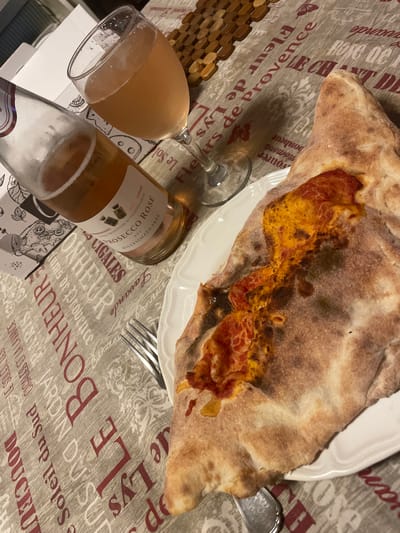 The image depicts a close-up view of a table setting, likely in a home or restaurant environment in Arese, Italy. A calzone, a folded pizza, is presented on a white plate. The calzone has a golden-brown crust and is topped with a visible layer of red sauce and melted cheese. Adjacent to the plate, a silver fork rests on a tablecloth. To the left of the calzone, a glass filled with a pale pink rosé wine sits beside a partially visible bottle of Prosecco Rosé. The bottle's label indicates it is "PROSECCO ROSE DOC MILLESIMATO 2025". The tablecloth is patterned with text and floral motifs in shades of grey and red. In the background, a wooden placemat or serving tray is visible. The overall impression is that of a meal or a celebratory drink. There are no people visible in the frame.