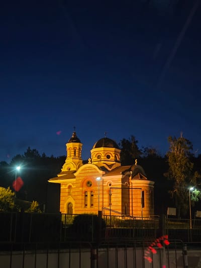 The image depicts a brightly illuminated Serbian Orthodox church at dusk or night in Dimovo, Bulgaria. The church has a distinct architectural style with multiple domes and is constructed with stone and brick, featuring orange and white horizontal banding. The main dome is larger and round, while a smaller, octagonal bell tower with a cross stands to its left. Exterior lighting casts a warm yellow glow on the building and its surroundings.

In the foreground, a dark metal fence runs horizontally across the lower portion of the frame. Beyond the fence, a hedge or low-lying shrubbery is visible. The background consists of a dense line of dark trees, silhouetted against the deep blue night sky. Two streetlights are visible; one to the left of the church, casting a bright beam upwards, and another to the right. There are some red reflections or lights in the lower part of the image, possibly from a vehicle or other light source, creating streaks across the fence. The sky is clear and dark, indicating it is either late evening or early morning. There are no people visible in the scene.