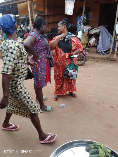 Three women are in an outdoor setting, possibly a market. The woman on the left is walking away, wearing a patterned dress and pink sandals. The woman in the center is standing with her hands on her hips, wearing a blue and red floral dress and green sandals. To her right, the third woman is holding a bag and a phone, wearing a red floral dress with a black panel and red sandals. Some goods are visible in the background.