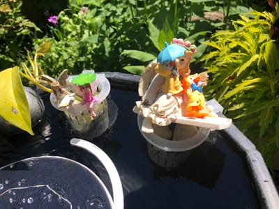 The image depicts a close-up outdoor scene, likely in a garden or patio setting in Severn, Canada. In the foreground, a black basin or birdbath contains dark water, reflecting the surroundings. A solar-powered fountain component is partially submerged in the water on the left, showing a circular dark panel with visible lines and some small water droplets and bubbles on its surface. A white, curved handle or support structure is also visible, extending from the right side of the solar fountain.

Further back in the basin, two small decorative containers hold miniature figurines. The left container holds a yellow leaf and what appears to be plant stems, with a small figurine of a fairy with brown hair and wings seated on the edge. A green lotus-like leaf is positioned above this fairy. The right container holds rocks and features a larger figurine of what looks like an angel or a child with blonde hair, holding a blue umbrella over a smaller figurine of a yellow bird-like creature wearing a blue bow tie. Another small orange figurine is also present.

The background is filled with lush green foliage and some small pink flowers, suggesting a vibrant garden environment. The lighting indicates it is a sunny day.