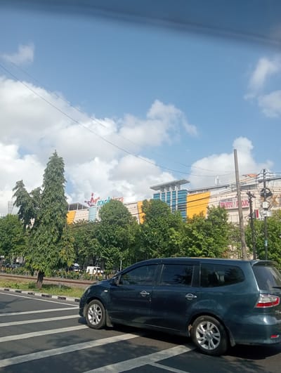 In Surabaya, East Java, a dark blue minivan is stopped at a pedestrian crossing in front of a large, modern commercial building complex. The building, partially obscured by trees, features a prominent yellow facade and a sign displaying "Royal Plaza". Another sign visible on the building reads "Mandiri". Tall evergreen trees and a variety of deciduous trees line the street and the building's perimeter. Overhead, power lines crisscross the clear blue sky, which is dotted with fluffy white clouds. The scene appears to be during daylight hours.