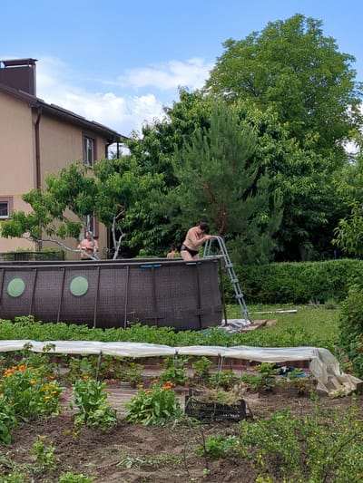 In a residential backyard in Boryspil, Ukraine, an above-ground swimming pool with a brown, woven texture is set up on a grassy lawn. A metal ladder leans against the side of the pool, and two individuals are visible near the pool. One person, wearing a black bikini, is climbing out of the pool using the ladder. Another individual, with dark hair pulled back, is partially visible inside the pool. In the background, a light-colored house with a brown roof and chimney stands adjacent to the pool. Lush green trees and bushes surround the property, and a garden area with young plants covered by a white tarp is situated in the foreground. The sky is partly cloudy.