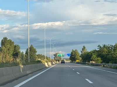 The image captures a highway scene, presumably near Urbnisi, Georgia. The perspective is from inside a vehicle, looking forward on a multi-lane asphalt road. The road is marked with white lines, indicating lanes of travel. On the left side of the road, there's a concrete barrier and then a grassy embankment with trees and vegetation. On the right, there's a metal guardrail and more trees. Several tall streetlights are visible along the left side of the road. In the distance, ahead on the highway, there are green and blue road signs, one of which appears to indicate directions with icons and text. A dark-colored car is traveling in the same direction, ahead of the viewer. The sky above is partly cloudy with dramatic cloud formations, suggesting either early morning or late afternoon lighting. The overall environment is an open, developed roadway with natural elements on the sides.