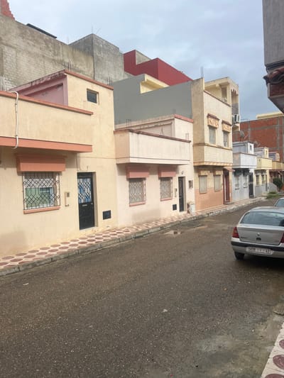 This image depicts a residential street in Ksar-el-Kebir, Morocco, under an overcast grey sky. The asphalt road in the foreground and mid-ground is wet with visible puddles, indicating recent precipitation. A sidewalk paved with alternating reddish-brown and beige circular tiles runs along the left side of the street, separated by a concrete curb.

A row of multi-story buildings lines the left side of the street. The nearest building is two stories, painted light beige with peach-colored trim, featuring a dark main entrance door with black decorative metalwork and two ground-floor windows equipped with security grilles. A white conduit runs vertically on its left facade. Adjacent buildings continue this architectural style, with varied heights, stucco finishes in shades of beige, cream, and light pink, and similar dark doors and grilled windows. Some buildings exhibit balconies or terraced sections. Upper levels and rooftops show exposed concrete blocks and rebar, suggesting ongoing construction or capacity for future expansion.

Further down the street on the right, a silver Renault Clio sedan is parked, facing away from the viewer. Its rear license plate reads "5898 | 1 | 42". Other buildings recede into the distance, some featuring satellite dishes and varying stages of completion with visible red, grey, and yellow painted sections. No individuals are present in the scene.