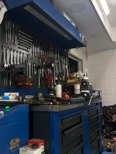 The image shows a cluttered workbench in a workshop setting. The workbench is blue and covered with various tools including screwdrivers, wrenches, pliers, and a drill. There are also cans, boxes, and other miscellaneous items on the workbench. The background is out of focus, but appears to be a garage or workshop with white tiled walls. The location is likely in a garage in Rakovica Urban Municipality, Serbia.