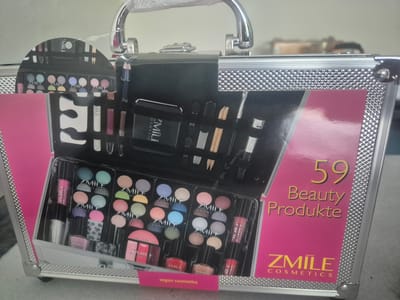 This is a medium shot, eye-level photograph of a silver and black makeup case. The case is open, revealing a variety of makeup products. The products are arranged in compartments within the case. Some of the products visible include eyeshadow palettes, lipsticks, nail polish, and brushes. The makeup case itself appears to be made of metal and has a handle at the top. The exterior of the case has a textured silver trim and a pink panel on the right side that reads "59 Beauty Produkte ZMILE COSMETICS". A small pink label on the bottom of the case reads "vegan cosmetics". The background of the image is blurred, but appears to be an indoor setting. There are no people visible in the image. The lighting is bright, suggesting it may be daytime or under artificial light. The overall impression is that of a retail product display.