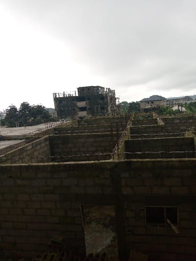 The image depicts a static scene of urban development and ongoing construction within an urbanized area of Buea, Cameroon, under an overcast sky.
In the immediate foreground, the lower level of a building is visible, constructed from grey concrete blocks. This structure features an open, unplastered doorway on the left and an open rectangular window aperture on the right, with a dark wooden plank leaning against its lower edge. Rebar extends vertically from the top of this foreground wall. Below this wall, at the base of the frame, fragmented views of construction materials are present.
The midground is dominated by multiple parallel and perpendicular concrete block structures, forming a grid of rectangular compartments. These appear to be the foundations or a partially completed ground floor slab of a multi-story building. Vertical rebar protrudes from the tops of these midground walls, indicating further vertical construction is planned or underway.
In the left background, a prominent, multi-story concrete building, appearing dark and unfinished or abandoned, rises above the midground structures. This building displays multiple window openings and exposed structural elements. To its left, several large, dense green trees are visible.
The right background reveals a more settled urban landscape, with various residential or commercial buildings featuring dark roofs, interspersed with dense green vegetation. These structures are situated on what appears to be sloping terrain, with distant hills faintly visible against the cloudy sky.
No individuals, vehicles, or active construction equipment are present in the frame. The scene conveys a state of architectural development and expansion.