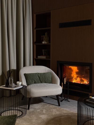 The image depicts an indoor scene, likely a living room, with a fireplace as the focal point. To the left, a light-colored armchair with a green pillow is placed near a side table. The side table holds a glass of water, a diffuser with reeds, and a small white bowl. Behind the armchair and side table, grey curtains are drawn. A bookshelf filled with various items, including a statue and books, is visible on the left wall. The fireplace on the right wall is lit, with flames and glowing embers visible. Next to the fireplace, a set of fireplace tools stands on the floor. A wooden wall paneling is present on the right side of the room. The floor is covered with a light-colored rug. The overall ambiance is warm and cozy. The location is identified as Bakovići, Montenegro.