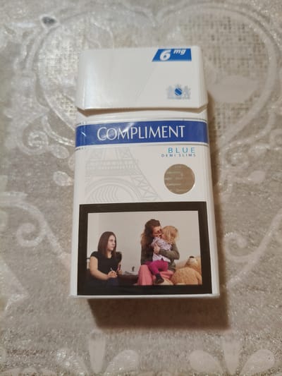 An overhead shot captures a pack of "Compliment Blue Demi Slims" cigarettes resting on a textured white surface. The pack is predominantly white with a blue band across the top, featuring the brand name in white capital letters. Below the brand name, a graphic of the Eiffel Tower is subtly imprinted in light grey. The bottom portion of the pack displays a photograph framed in black, depicting two women and a child. The woman on the left, wearing a black top, is looking to her right, holding a pen. To her right, another woman in a green sweater and pink pants holds a young child, who is reaching out. A teddy bear sits to the right of the women and child. The image does not contain any information that would suggest its location in Chornobaivka, Ukraine, or any other specific city.