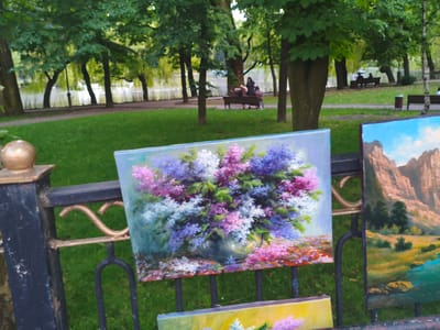 In Verkhnii Lars, Russia, a park setting is depicted with lush green grass and numerous trees, some with weeping branches arching over a body of water. In the background, several people are visible sitting on benches. In the foreground, two paintings are displayed on a black metal fence. The painting on the left is a still life of a large bouquet of lilacs in a vase, with a scattered arrangement of smaller flowers on a surface below. The painting on the right is a landscape featuring a river flowing through a valley with red-brown rock formations in the background and scattered trees. The scene appears to be an outdoor art display in a public park.