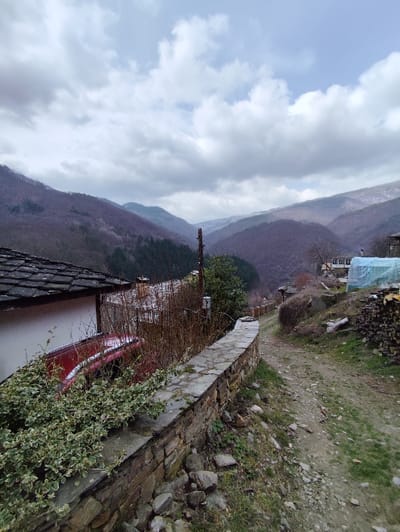 This image depicts a mountainous rural landscape within the city of Kosovo, Bulgaria, under a heavily overcast sky composed of gray and white clouds. In the immediate foreground, a low, curving dry-stone wall, constructed from irregular flat stones, is partially covered by green and white variegated ivy. To the left of the wall, a segment of a red vehicle is visible, partially obscured by bare shrubbery and ivy, situated next to a building with a dark, possibly slate, roof and light-colored walls. To the right of the stone wall, an unpaved, uneven dirt path with scattered rocks and patches of green grass extends into the midground. Further along the path on the right, a slope supports sparse vegetation and a stacked pile of cut firewood. In the midground, additional traditional buildings with dark roofs are discernible, one of which has a light blue tarp draped over a section. A wooden utility pole with visible wires stands on the left side of the path. The background features successive ranges of hills and mountains, largely covered by bare deciduous trees, indicative of a colder season. No individuals, actions, or direct interactions are present in the scene.