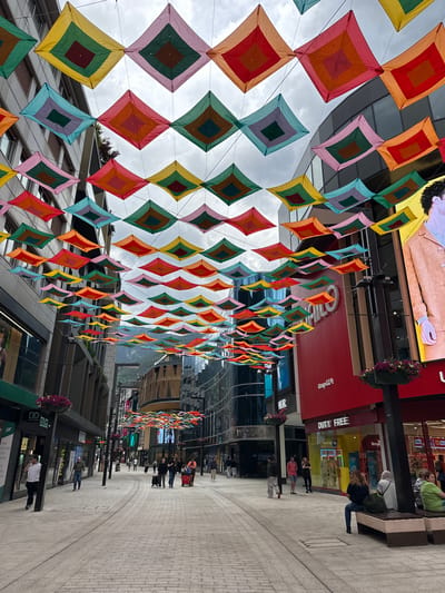 This image captures a vibrant pedestrian shopping street in Andorra la Vella, Andorra, adorned with numerous colorful diamond-shaped fabric decorations suspended overhead. These banners, featuring concentric squares in diverse hues such as red, orange, yellow, green, blue, and pink, create a striking canopy against the overcast daytime sky.
The street is flanked by modern, multi-story buildings housing various retail establishments. On the right side, prominent signage includes "DUTY FREE", "GrupCCA", and "DOUGLAS". Other discernible texts further down this side read "TAPUGEM", "LET'S", "Winsto", and "West". On the left, a sign for "AMORABAN" and "DOUGLAS" is visible, alongside smaller text like "Servei" and "tota". A sign with "EL SOU" is also present.
Several people are seen throughout the scene, engaged in everyday activities such as walking, standing, or sitting on a public bench. The setting suggests a bustling commercial area, characterized by its modern architecture and the festive, artistic street decorations. The weather is overcast, but the bright colors of the decorations maintain a cheerful and lively atmosphere.
