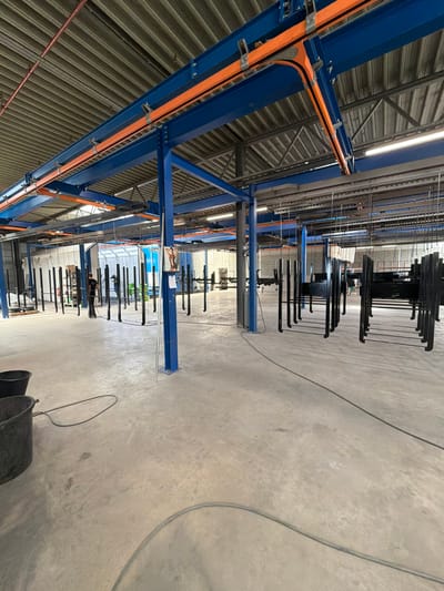 The image shows an indoor industrial setting, likely a factory or workshop, in Mol, Belgium. The scene is dominated by a large blue metal structure, possibly part of an overhead conveyor system or racking. Orange conduits are attached to this structure, running along its beams. The ceiling is corrugated metal, and fluorescent lighting is visible.
Several people are present, appearing to be workers. One individual is visible near the middle of the frame, wearing dark clothing and working on or near the overhead system. Another person, also in dark clothing, is further down and partially obscured by hanging objects.
The primary activity appears to be some form of manufacturing or processing, suggested by the hanging metal parts. These parts, which look like frames or components, are suspended from the overhead system. They are dark in color and appear to be awaiting treatment or assembly. The concrete floor is clean, with some electrical cables running across it.
The time of day is not clearly discernible, but the artificial lighting suggests it's an indoor operation. The weather is irrelevant as the scene is indoors. The overall impression is one of an active industrial environment. No visible text is present in the image.
