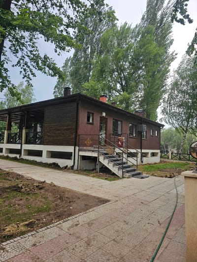 The image shows a brown building with a covered patio area and a white foundation. A set of concrete stairs with metal railings leads up to the entrance of the building. The building is surrounded by green trees and a cloudy sky. The ground in front of the building is a paved walkway with some patches of dirt and grass. A green hose is visible running across the walkway. The location is identified as Chong-Aryk, Kyrgyzstan. There are no people visible in the image.