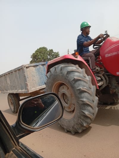 A red tractor with a partially visible "Mahindra" logo is observed moving right-to-left on a light brown paved road in Peyer, Nigeria. It is pulling a rectangular, open-top, blue-grey metal trailer. The trailer appears worn and dirty. A male driver, with dark skin, wearing a dark blue short-sleeved shirt, dark pants with light stripes, and a green hard hat, operates the tractor, gripping the steering wheel and looking forward. The tractor's large, knobby rear right tire is prominent. The scene is captured from the perspective of an adjacent vehicle, with its black side mirror visible in the lower left foreground, reflecting a partial image of an arm/hand and an interior surface. The background features a light sky and a green-leafed tree.