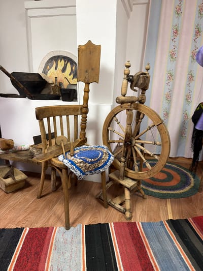 This image depicts a room with traditional elements, possibly a historical exhibit or a well-preserved living space. In the background, a white wall features a decorative rendering of a fireplace with flames, suggesting a symbolic representation of warmth or hearth. To the right, pastel-colored curtains with floral stripes hang, adding a touch of decorative softness.

The foreground is dominated by antique wooden objects. A rustic wooden spinning wheel stands prominently, its intricate structure suggesting a bygone era of textile production. Beside it, a simple wooden chair is placed. On the chair rests a small, round rug with a blue and white patterned center and a brown and white outer edge, possibly handmade. A wooden board with what appear to be spinning tools lies across the chair.

To the left of the chair, a wooden bench holds various items, including a dark pot or pan with a handle, a folded cloth, and some smaller tools. A wooden box with a handle is visible on the floor beneath the bench.

Two colorful woven rugs are spread on the wooden floor, one in the foreground with bold stripes of black, red, orange, and white, and another circular rug with concentric rings of green, orange, and purple further back. A hint of a person wearing a purple garment and a black head covering with fringe is visible in the far right background, partially obscured by the curtains.

The lighting is even and diffuse, illuminating the scene without harsh shadows, suggesting an indoor setting under soft light. The overall impression is one of a curated display of traditional domestic tools and decor, evoking a sense of history and cultural heritage. The specific location in the city of Ust'-Kabyrza, Russia, is not visually apparent from the image itself, but the objects suggest a connection to rural or folk traditions.