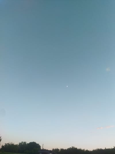 The image depicts a wide expanse of a pale blue sky, transitioning to a lighter hue near the horizon. A crescent moon is visible in the upper right quadrant of the sky. Below the sky, a dense line of dark green trees forms the horizon. To the right of the trees, a small portion of a grey structure with a dark roof is partially visible. The scene appears to be an outdoor landscape in the city of Sosnivka, Ukraine, possibly taken during twilight due to the soft lighting and visible moon.