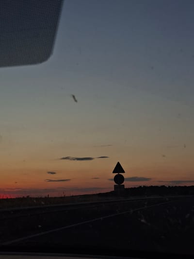 The image depicts a sunset sky transitioning from blue-grey to orange-red with some scattered clouds. A triangular arrow traffic sign with a circular attachment is visible, and what appears to be a railroad track or road runs alongside a line of trees. A screen is in the bottom left corner. It is not possible to confirm that the scene is located in Momkovo, Bulgaria.