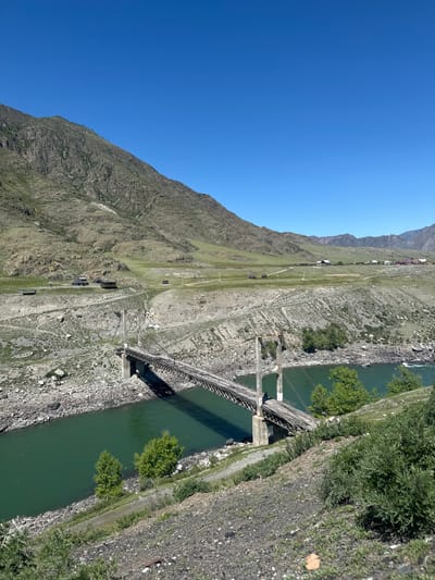 The image shows a bridge crossing a river in Inya, Russia. The bridge is a suspension bridge made of concrete and metal. It spans across a river with rocky banks. The river is green in color. On one side of the river, there is a steep mountain with some sparse vegetation. On the other side, there is more dense vegetation, including trees and bushes. The sky is clear and blue. There are a few buildings visible on the mountain side near the bridge.
