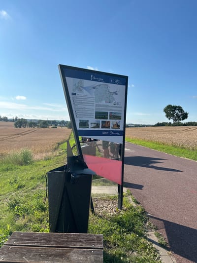An informational sign with a map and text is positioned next to a paved path on a sunny day. The sign has a dark blue background with white text and graphics, and it is supported by a black metal frame. A portion of a red bench is visible in the foreground, to the left of the sign. A black trash can, partially filled with black plastic bags, sits behind the red bench. The path appears to be a bike or walking trail and is bordered by green grass and a field of golden crops. In the background, trees and a clear blue sky with scattered clouds are visible. The image appears to be taken in Bałdowo, Poland, indicated by the signage. No people are clearly visible.