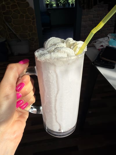 A hand with bright pink fingernails holds a tall glass mug filled with a thick, white milkshake topped with whipped cream. A yellow plastic straw extends from the whipped cream. The mug sits on a dark surface, possibly a table, with a shadow cast across it. In the background, a textured wall, a doorway with plants visible through it, and a brick wall can be seen. The lighting suggests it is daytime, with sunlight coming from the left, creating highlights and shadows. The scene is indoors, likely in a home or a cafe. There is no indication of the city of Ivanovka, Russia, or any specific location.