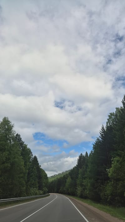 The image shows a two-lane asphalt road curving through a dense forest. The road has white dashed lines marking the lanes and a metal guardrail on the left side. The trees are tall and green, creating a tunnel-like effect. The sky is partly cloudy with patches of blue visible. In the distance, rolling hills covered in trees are visible. The scene appears to be a natural landscape in a rural or mountainous area. There are no people or specific buildings visible in the image. The location is identified as Staroye Tataurovo, Russia, suggesting a remote setting within Russia.