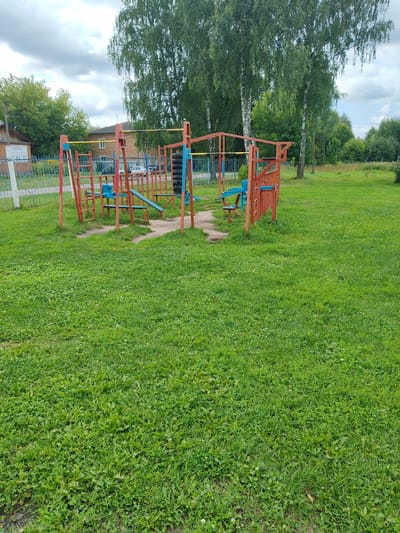 In Drezna, Russia, a public outdoor exercise apparatus stands on a well-maintained green lawn. The apparatus consists of red metal posts with blue seats and yellow horizontal bars, featuring elements like a pull-up bar and what appear to be leg press or sit-up benches. A stack of tires is mounted on the side of the structure. Behind the exercise equipment, a metal fence encloses a paved area with some buildings and trees visible in the background. The sky is overcast with scattered clouds, and the overall environment suggests a park or a communal recreational space.