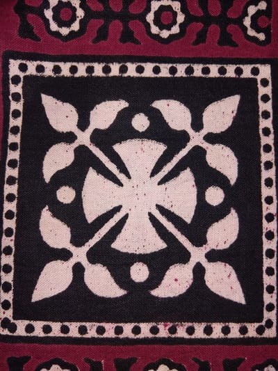 The image shows a close-up of fabric with a repeating pattern. The central design is a square with a black background, outlined by a border of white dots on a white background. Inside the black square, a stylized white design resembles a cross with four leaf-like shapes radiating from the center, each with a small white circle between them. The fabric appears to be a natural material, possibly cotton, with visible texture and small red speckles on the white areas. To the left and right of the central square, the fabric has a maroon background with a black and white pattern of floral or geometric motifs. The overall impression is of a decorative textile, possibly part of a garment, tapestry, or home furnishing. The lighting is consistent, suggesting an indoor setting. No people, animals, or specific activities are depicted. No text is visible. The style of the pattern might suggest traditional South Asian textile art. Given the provided location context, it is plausible this fabric is from Haveli Jhangranwali, Pakistan.