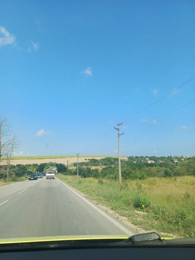 The image shows a view from a vehicle driving on a road. The road is paved and has white lane markings. There are several cars on the road ahead. The surrounding environment is rural, with fields and trees. In the distance, there is a village with houses. The sky is blue with a few scattered clouds. The photo appears to have been taken in Kumanovo, Bulgaria.
