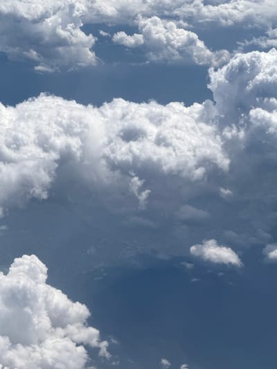 The image is an aerial view of clouds and the Earth's surface, taken from an airplane. The clouds are dense and puffy, with varying shades of white and gray, indicating sunlight filtering through them. Beneath the clouds, the Earth's surface appears as a dark, textured expanse, possibly mountainous terrain or a large body of water. There are no people or man-made structures clearly visible in the image, suggesting a remote or natural landscape. The location is identified as Nanahar, India.