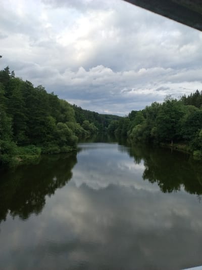 The image shows a wide, calm river flowing through a densely forested area. The trees on either side of the river are lush and green, with their reflections clearly visible on the water's surface. The sky is overcast with dramatic clouds, allowing some light to filter through. The perspective suggests the photo was taken from a bridge or elevated position looking downstream. The scene is serene and natural, with no signs of human activity or man-made structures except for what appears to be a partial view of a dark railing in the upper right corner, possibly indicating the vantage point. This location is identified as the Vltava River near Stádlec, Czechia.