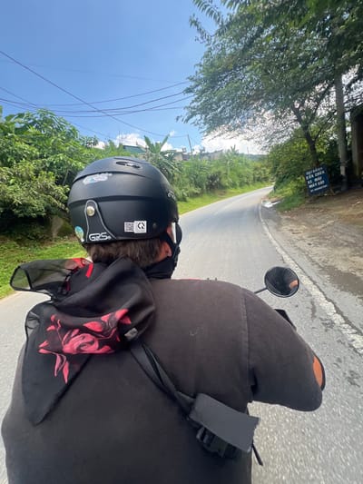 The image shows a person riding a motorcycle on a road in Bình Lư, Vietnam. The person is wearing a black helmet and a dark jacket with a red and black floral scarf draped over their shoulders. The road is paved and curves to the left, with lush green vegetation and trees lining both sides. In the distance, there are some buildings and utility poles with wires. A blue sign is visible on the right side of the road, displaying Vietnamese text. The sky is blue with some white clouds. The overall scene depicts a travel or commute through a rural or semi-rural area.