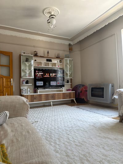 The image depicts the interior of a living room, likely a home, situated in Körfez, Turkey. The scene is well-lit and appears to be in daylight, with natural light entering from an unseen window.
Dominating the central area is a modern media console with a large flat-screen television displaying the YouTube interface. The console features a mix of wooden drawers and open shelving, with glass-fronted cabinets on either side of the TV, displaying decorative items. Above the television, the wall is adorned with various decorative objects and figurines, including what appears to be a miniature mosque or tower and a sailing ship model.
To the left of the media console, a closed wooden door with a vertical handle is visible. The wall above the console and along the ceiling is decorated with intricate molding. A decorative ceiling light fixture with three glass shades hangs from the ceiling.
On the right side of the room, a wall-mounted gas heater is prominent, with a visible gas pipe connection. Adjacent to the heater is a circular decorative wall piece featuring a silhouetted couple with balloons against a cosmic background.
The floor is covered by a large, plush, off-white carpet, with a smaller, patterned rug placed in front of the gas heater. A portion of a light-colored sofa with a textured fabric and a throw blanket is visible in the foreground on the left. Another part of a sofa, with a blanket draped over it, is partially visible on the right. The walls are painted in a neutral, light beige color. The overall impression is of a comfortably furnished and decorated living space.