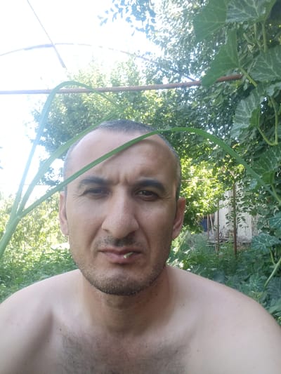 This is a close-up, selfie-style photograph taken outdoors in a rural or semi-rural setting, possibly in Karaganda, Uzbekistan. The subject is a man with short, dark hair and a light beard, looking directly at the camera. He is shirtless, revealing his shoulders and upper chest, which show some sparse body hair. A piece of green vegetation is visible in his mouth, held between his teeth. The background consists of lush green foliage, including large, broad leaves that appear to be part of a vine or gourd plant, and other trees and plants. A simple structure, possibly an outdoor frame or trellis, is visible in the upper portion of the image, along with what appears to be a portion of a building in the distance. The lighting suggests it is daytime, with sunlight filtering through the leaves, creating dappled light and shadows. The overall impression is of a casual, candid moment captured in a natural environment.