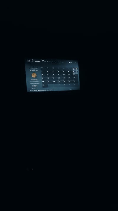 The image shows a screen displaying a calendar interface. The calendar is for Saturday, May 2nd, 2020, and lists "EFL League Two" with a Cambridge crest. Below this, it shows "Cambridge" and then "60 days until the opening." The calendar grid shows dates from the 26th of April to the 6th of June. The interface has menu options at the bottom, including "Events," "Home," "Settings," "Help," and "Menu." The image is dark, suggesting it might be taken in a low-light environment or is a screenshot of a dark-themed application. There is no indication of the city of Zhanadaur, Kazakhstan, or any people in the image.