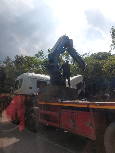 In Kisanga, Tanzania, an outdoor scene depicts a recovery operation on an asphalt road. A red flatbed truck with visible storage compartments and two rear axles is parked, equipped with a blue hydraulic crane mounted on its rear section. The crane, bearing the text "TEL: 0783544158," is extended and articulated towards the right side of the frame. One individual, dressed in dark clothing, stands on the flatbed near the crane's base, facing away from the camera, seemingly operating or overseeing the crane's function. A white truck cab, possibly detached from its chassis, is partially visible off the road to the right, situated amongst dense green foliage. An indistinct object or figure is discernible on the ground near the white truck cab. The sky is partly cloudy with significant bright sunlight, and a dense treeline forms the background.