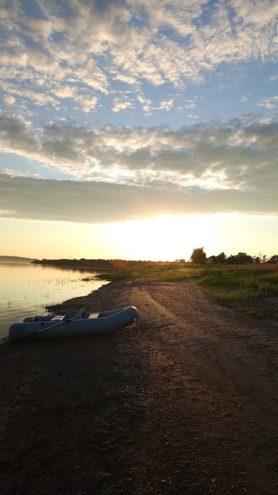 The image depicts an outdoor landscape at a body of water, located in Шолья, Russia, during the golden hour, indicating either sunrise or sunset. The sun is positioned low on the horizon in the central-right portion of the frame, emitting intense golden light that illuminates the scene and creates prominent long shadows. The sky above features scattered altocumulus clouds, backlit by the sun, displaying hues of white, grey, and soft orange against a pale blue background.
In the foreground, a dark dirt and gravel path with discernible tire tracks extends along the shoreline from the lower right towards the middle distance. An inflatable grey boat, equipped with two oars, is beached on the pebbly shore in the lower-left quadrant of the image. A distinctive logo, resembling a stylized 'S' or 'D' with a wavy line, is visible on the bow of the boat. The calm water surface, indicative of a lake or wide river, subtly reflects the ambient light and sky. Sparse vegetation, consistent with reeds or grasses, grows along the immediate waterline. Beyond the dirt path, an expanse of green grass and foliage transitions into a distant treeline that forms the horizon on the land side. No human or animal figures are present within the scene.