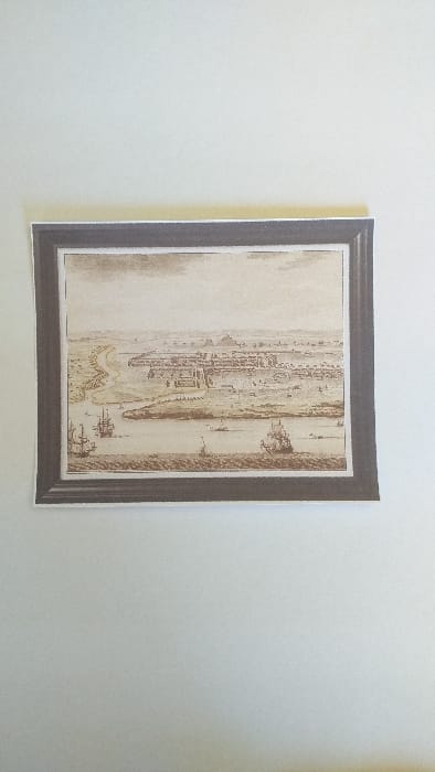 The image displays a photograph of a sepia-toned historical print, presented within a dark, simulated frame against a plain, light-colored background. The print itself depicts an elevated, panoramic view of the city of Limpok (Jambi), Indonesia.

The scene features a wide river dominating the mid-ground, containing a large elongated island or peninsula. Numerous sailing vessels are visible on the water, including two prominent multi-masted ships positioned in the lower-left portion of the river, and several smaller boats scattered across the waterway, indicating active maritime traffic. The city of Limpok is situated on the riverbank, characterized by a dense arrangement of structures, some of which appear as fortified walls or large building complexes. The architecture suggests a historical period. The background shows distant, undefined terrain under a light sky, while the immediate foreground consists of a strip of land bordering the river. No individual human figures are discernible within the detailed illustration.