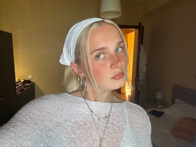 The media file is a portrait of a young woman with blonde hair, wearing a white translucent top and a white headscarf. She has a fair complexion with freckles across her nose and cheeks, and she is wearing lip gloss. She has large hoop earrings and a nose ring. The image is oriented upside down. The woman's face is the primary focus. In the background, a light-colored wall and a dark-colored surface, possibly a piece of furniture, are visible. There is also a window with warm lighting emanating from it, and what appears to be a white ceramic cup or planter on a surface. The lighting suggests it might be indoors, possibly in a room with natural light coming from a window or artificial lighting. The overall mood seems relaxed and casual. No text is visible in the image.