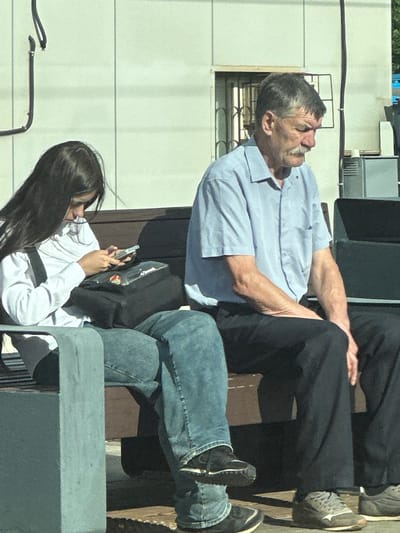 The image shows two people outdoors, possibly in a public space in Lobnya, Russia. The subject in the foreground, a woman with long dark hair, is wearing a white shirt and jeans and is looking at her phone. The second subject, an older man with graying hair and a mustache, is wearing a light blue collared shirt and dark pants. He is looking upwards with a slightly pained expression. The scene appears to be set during the daytime, with natural lighting. The background shows a building with a light-colored facade and some architectural details like windows and railings. There are also other people partially visible in the background, suggesting a populated area. The overall impression is a candid moment captured between two individuals, with one focused on their device and the other looking away.